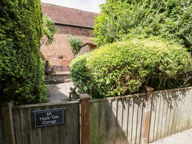 A garden area with dense shrubbery and a wooden fence with a sign reading Hock-Tide Cottage at Kenilworth