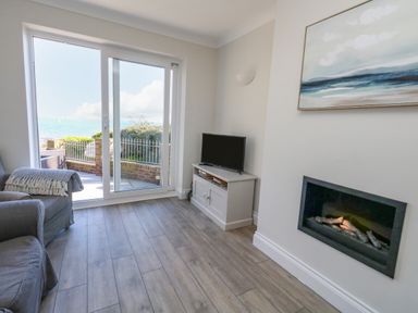 A living room with a gray armchair a small TV on a cabinet a wall-mounted fireplace and sliding glass doors leading outside at Castle Cove Cottage in Weymouth