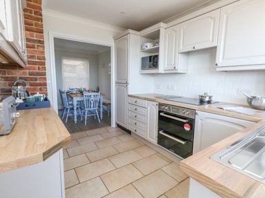A kitchen with white cabinets wooden countertops a built-in oven a toaster and a dining area with a wooden table and blue chairs at Castle Cove Cottage in Weymouth