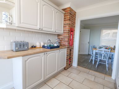 A kitchen with white cabinets wooden countertop a toaster and a tray with teapot and cups next to a red brick wall with a red mailbox leading to a dining room with a wooden table and blue chairs at Castle Cove Cottage Weymouth