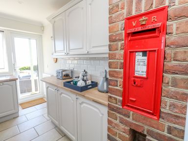 A kitchen with white cabinets and a wooden countertop with a tray of teapots and cups and a red post box mounted on a brick wall at Castle Cove Cottage in Weymouth