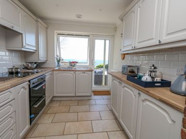 A kitchen with white cabinets wooden countertops a stove a toaster a kettle a French press and a door with a window showing a sea view at Castle Cove Cottage Weymouth