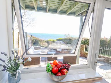 A kitchen window with a bowl of fruit and a vase of flowers overlooking an outdoor patio and the sea at Castle Cove Cottage in Weymouth