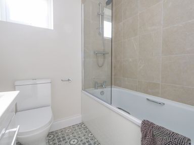 A bathroom with a toilet, bathtub with shower, glass screen, and patterned tile floor at Castle Cove Cottage in Weymouth