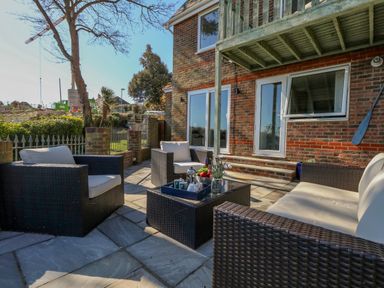 An outdoor patio with wicker furniture and cushions near a brick house with glass doors at Castle Cove Cottage in Weymouth