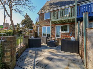 A patio area with outdoor seating and a table next to a brick house with a balcony at Castle Cove Cottage in Weymouth