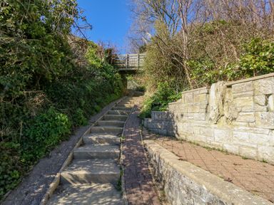 An outdoor stone staircase with a narrow path beside it leading under a small wooden bridge surrounded by greenery and stone walls at Castle Cove Cottage in Weymouth