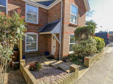 The exterior entrance of a brick house with a white door and windows surrounded by small garden beds with shrubs and plants at Castle Cove Cottage in Weymouth