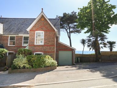 A brick house with a green garage door and bushes in front near trees and a view of the sea at Castle Cove Cottage in Weymouth