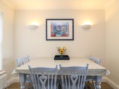 A dining room with a rectangular table covered by a polka dot tablecloth and four chairs with a framed picture and two wall lights on the wall at Castle Cove Cottage in Weymouth