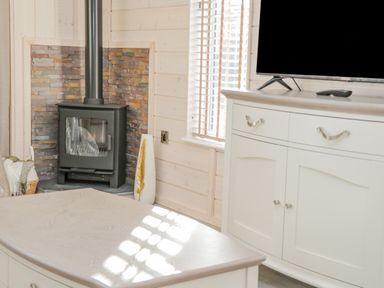 A corner with a black wood burning stove surrounded by stone tiles next to a window and a white cabinet with a TV on top at No 27 The Crab Pot in Runswick Bay near Staithes