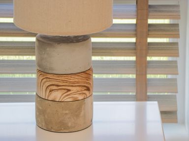 A table lamp with a beige shade and a base made of stacked concrete and wood blocks on a white surface by a window with striped blinds at No 27 The Crab Pot in Runswick Bay near Staithes