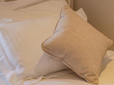 A close up of a beige textured cushion on white bed pillows and sheets in a bedroom