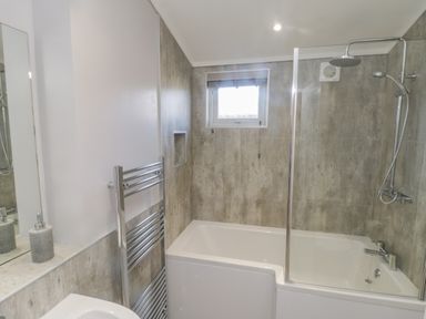 A bathroom with a bathtub with showerhead and glass screen a towel rack and a window at No 27 The Crab Pot in Runswick Bay near Staithes