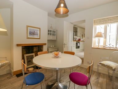 A dining room with a round table and colorful chairs next to a fireplace and window at Primrose Cottage in Winchcombe