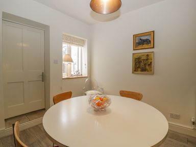 A dining area with a round white table and wooden chairs near a window with a lamp and two framed paintings on the wall at Primrose Cottage in Winchcombe