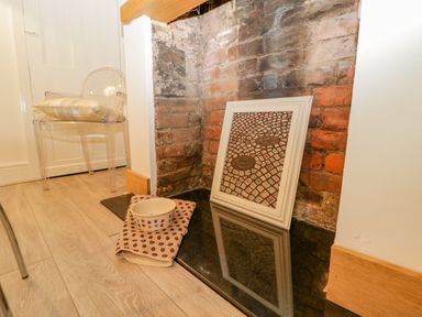 A corner with exposed brick wall containing a framed picture on the floor next to a pet bowl on a cloth and a transparent chair with a plaid cushion in a room at Primrose Cottage in Winchcombe