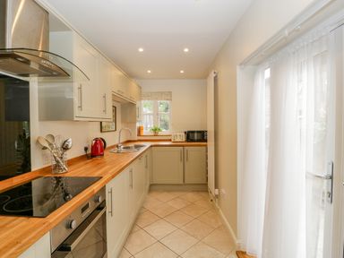 A kitchen with wooden countertops, cream cabinets, stove, oven, sink, toaster, kettle, microwave, window, and glass door with white curtains at Primrose Cottage Winchcombe
