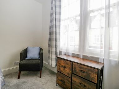 A corner of a room with a dark chair with a blue cushion and a wooden drawer unit by a window with checkered curtains at Primrose Cottage in Winchcombe