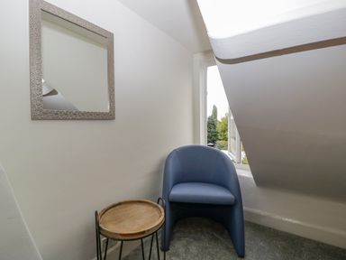 A small corner with a blue chair a round wooden side table a wall mirror and a window with a view of trees at Primrose Cottage in Winchcombe