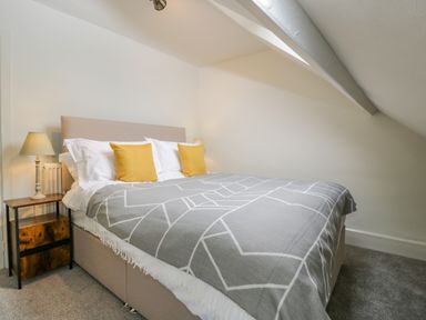 A bedroom with a bed with yellow pillows and a gray blanket beside a bedside table with a lamp at Primrose Cottage in Winchcombe
