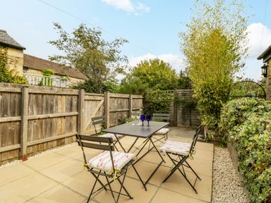 An outdoor patio with a table and four chairs with cushions surrounded by wooden fencing and plants at Primrose Cottage in Winchcombe
