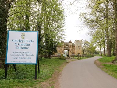 An entrance sign for Sudeley Castle and Gardens near a paved driveway leading to a stone gatehouse surrounded by trees