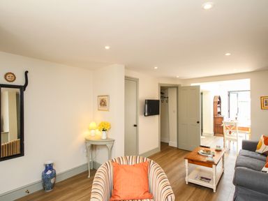 A living room with a striped armchair and orange pillow a small table with a lamp and flowers a coffee table with cups and plates a grey sofa and a wall-mounted television at Blanche Cottage in Brewers Quay Harbour