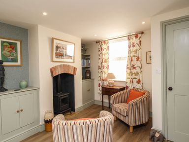 A sitting area with two striped armchairs a wood stove fireplace bookshelves a window with floral curtains and a side table with lamp at Blanche Cottage in Brewers Quay Harbour