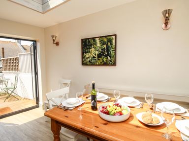 A dining room with a wooden table set with plates glasses a bottle of wine and a bowl of fruit with open doors leading to a small balcony at Blanche Cottage Brewers Quay Harbour
