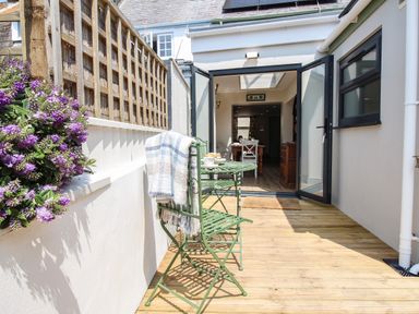 A small outdoor patio with green metal table and chairs a blanket on a chair flowers on the left and open doors leading inside at Blanche Cottage Brewers Quay Harbour