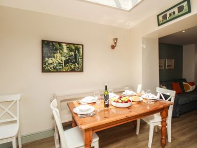 A dining area with a wooden table set with plates glasses a bottle of wine and a bowl of fruit surrounded by white chairs at Blanche Cottage Brewers Quay Harbour