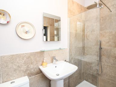 A bathroom with a pedestal sink, wall mirror, soap dispenser, toilet, shower area with glass partition and beige tiled walls at Blanche Cottage Brewers Quay Harbour