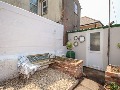An outdoor courtyard with a bench covered with a blanket and a cup on it next to raised brick flower beds and a white door at Blanche Cottage Brewers Quay Harbour