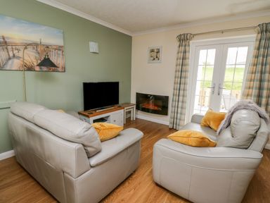 A living room with two leather sofas yellow cushions a television an electric fireplace and double doors with curtains at Oak Tree Cottage in Aberporth