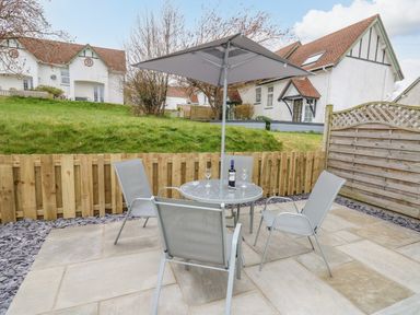A patio with a round glass table four chairs and an umbrella with houses and grass in the background at Oak Tree Cottage in Aberporth