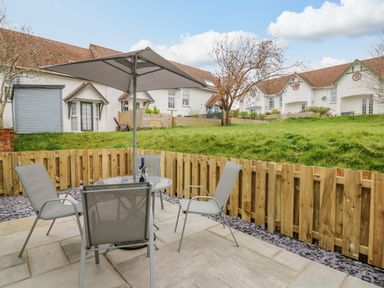 An outdoor patio with a glass table, four chairs, an umbrella, and a wooden fence overlooking grassy yards and white houses at Oak Tree Cottage in Aberporth