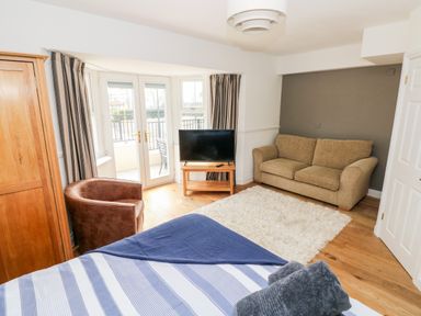 A bedroom with a striped bedspread a beige sofa a brown armchair a wooden wardrobe and a TV on a wooden stand at Puffins in Tenby