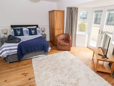 A bedroom with a bed side tables a wooden wardrobe a brown chair a white rug and a TV on a wooden stand with a door to a balcony at Puffins in Tenby