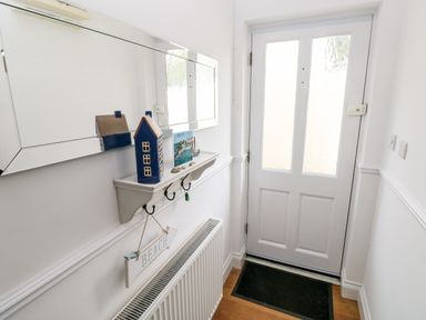 An entrance hallway with a white door a black doormat a wall shelf with hooks and decorative items and a large mirror above the shelf at Puffins in Tenby