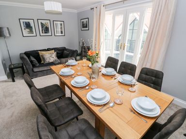A dining room with a wooden table set for eight and a sofa against a gray wall at Puffins in Tenby