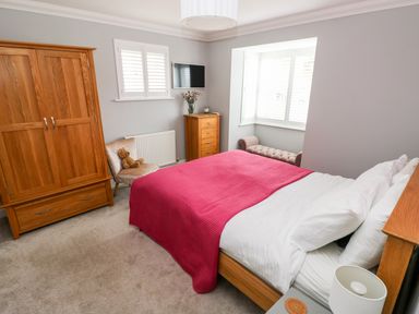 A bedroom with a double bed with white sheets and a red blanket a wooden wardrobe a wooden chest of drawers a chair with a teddy bear a wall-mounted TV and a window seat at Puffins in Tenby