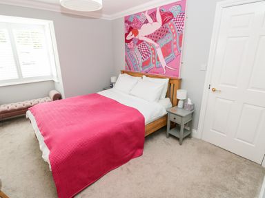 A bedroom with a bed covered by a pink blanket a wooden headboard a painting above the bed bedside tables with lamps and a window with white blinds at Puffins in Tenby