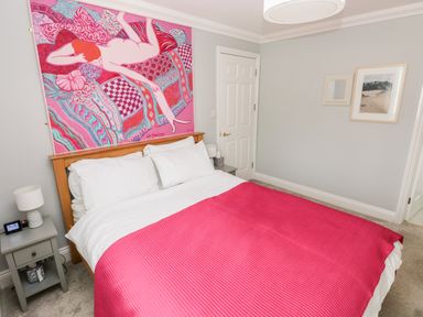 A bedroom with a bed covered with a pink blanket bedside tables with lamps a colorful painting above the bed and framed pictures on the wall at Puffins in Tenby