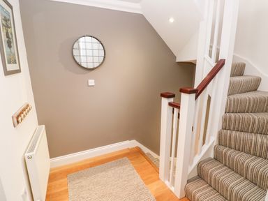 A staircase with striped carpet and white railings and a small hallway with wooden flooring and a round mirror on a grey wall at Puffins in Tenby