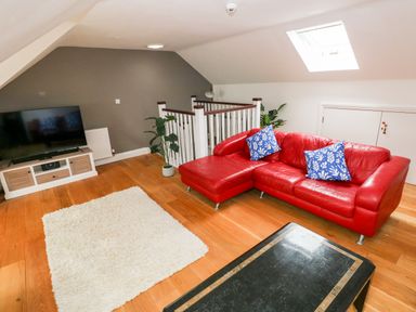 A living room with a red sectional sofa with blue pillows a television on a wooden stand a white rug wooden flooring and a skylight at Puffins in Tenby