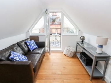 A small living area with a black leather sofa with blue patterned cushions a gray console table with a lamp and globe a pouf and a balcony door at Puffins in Tenby