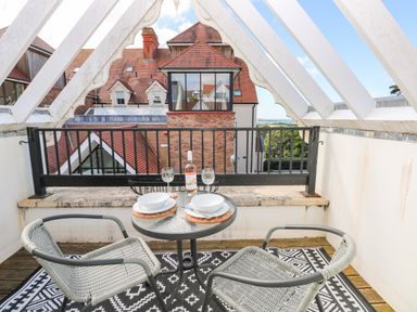 A balcony with two chairs a small table set for two with a bottle and two glasses overlooking tiled rooftops at Puffins in Tenby