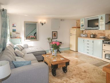 A living room with a grey sofa wooden coffee table and kitchen area with pastel cabinets at Nightingale Cottage in Lostwithiel