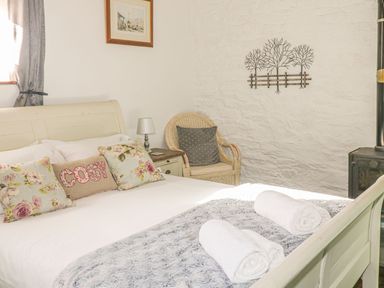 A bedroom with a bed with floral and cosy pillows a wicker chair bedside table with lamp and wall art at Nightingale Cottage in Lostwithiel